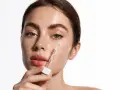 Kobieta aplikuje serum z pipetki na policzek. Skóra pełna piegów, nawilżona, z widocznym blaskiem. Kluczowe składniki: retinol i ceramidy.