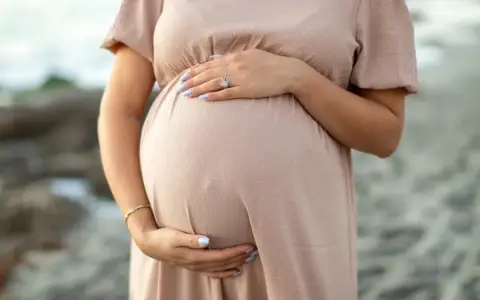 Brzuszek ciężarnej kobiety w beżowej sukience, dłonie delikatnie go obejmują. Warto pamiętać o bezpieczeństwie i unikać stosowania **retinolu w ciąży**.