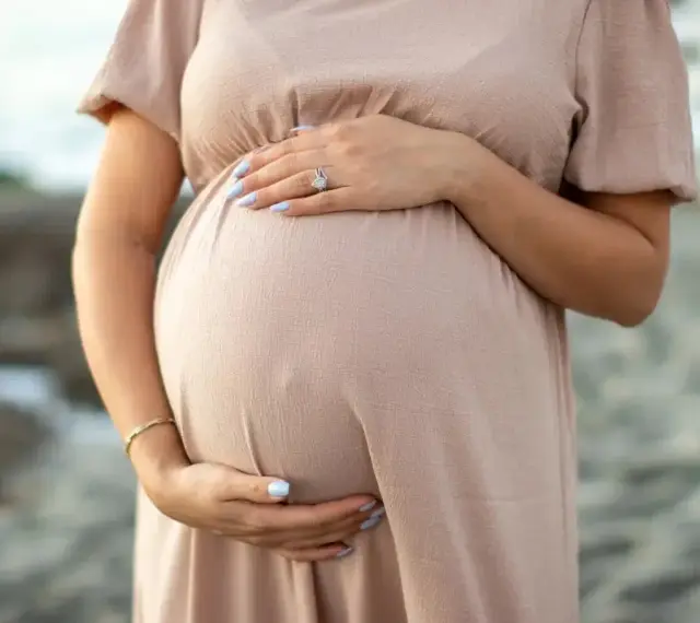 Brzuszek ciężarnej kobiety w beżowej sukience, dłonie delikatnie go obejmują. Warto pamiętać o bezpieczeństwie i unikać stosowania **retinolu w ciąży**.