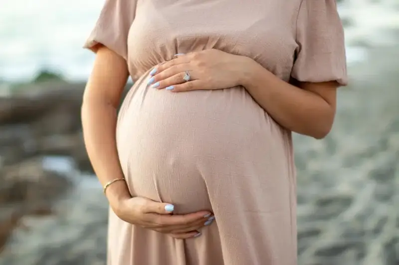 Brzuszek ciężarnej kobiety w beżowej sukience, dłonie delikatnie go obejmują. Warto pamiętać o bezpieczeństwie i unikać stosowania **retinolu w ciąży**.
